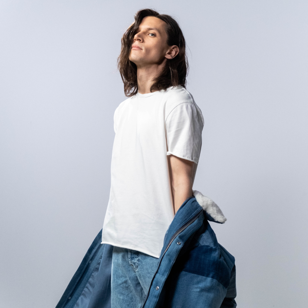 Person wearing a white t-shirt and blue jeans with a gray jacket over their shoulder on a light gray background